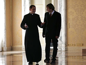 Andreas Englisch läuft an der Seite eines Priesters in einer marmornen Halle her. 