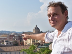 Andreas Englisch steht auf einer Erhöhung und deutet auf die Stadt Rom hinter ihm.