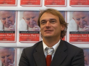 Andreas Englisch im Anzug vor einer Wand, auf der mehrmals das Cover seines Buches "Johannes Paul II" abgedruckt ist.